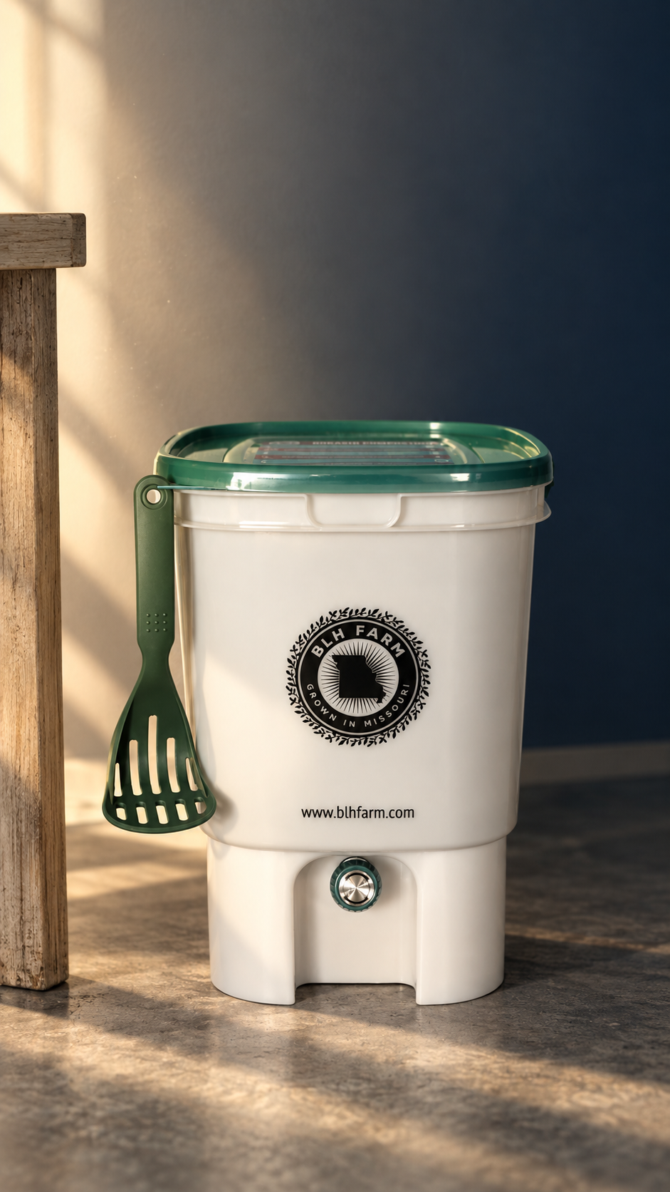 BLH Farm bokashi bin with green lid, slotted paddle and drain tap, on concrete beside an oak table